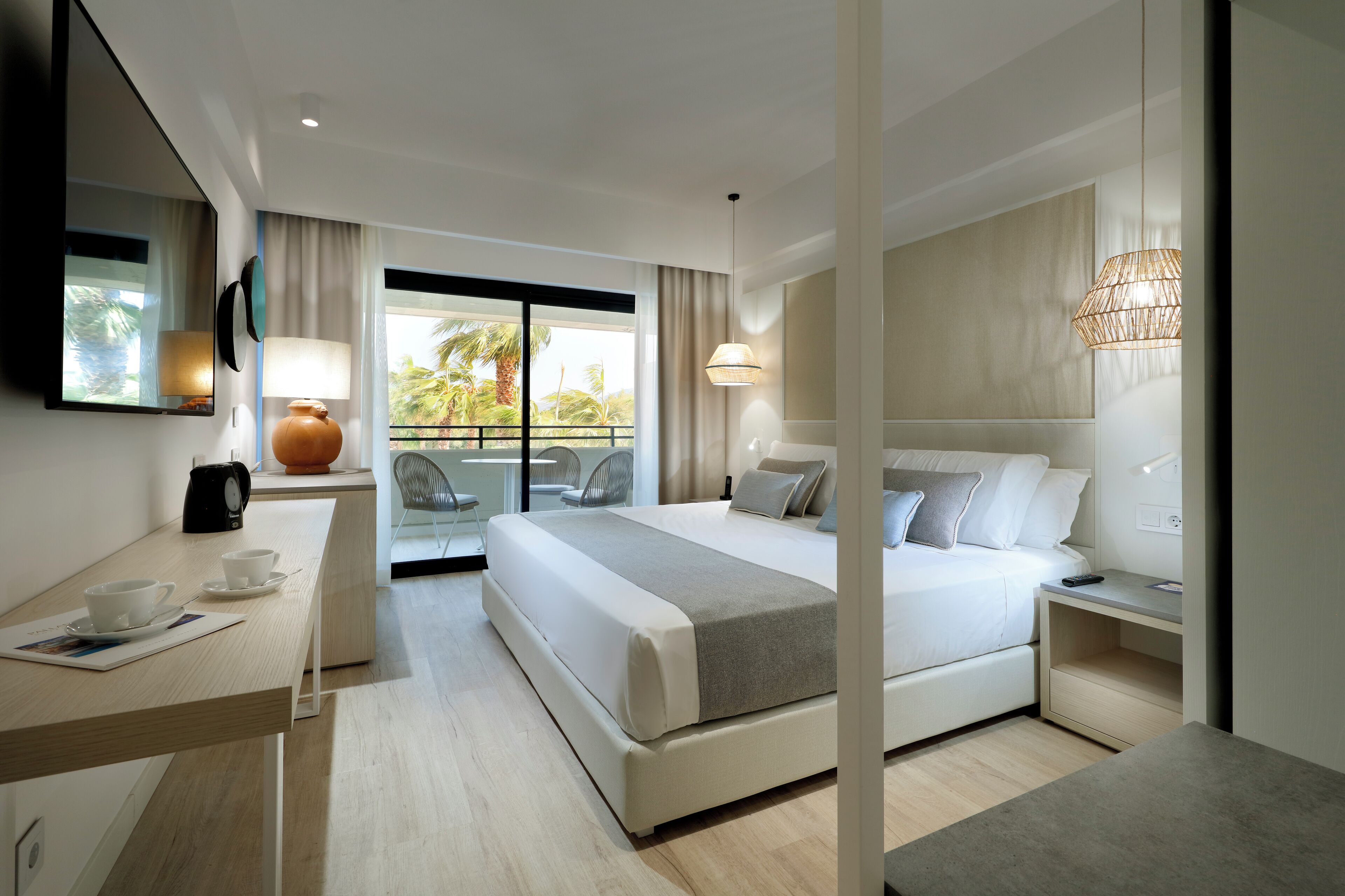 A modern hotel room with a large bed, a desk with a coffee maker, a chair, and a balcony with a view of palm trees.