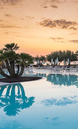 A poolside area with lounge chairs and umbrellas, palm trees, and a sunset sky.