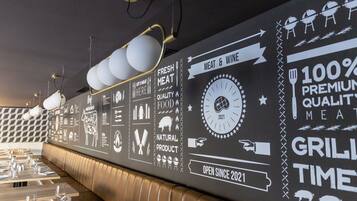 A restaurant interior with a long counter, dining tables, and a wall decorated with food and grill-related graphics.
