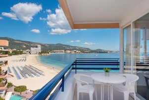 Un balcón con vistas a la playa y al mar, sillas blancas y una pequeña mesa con una planta.