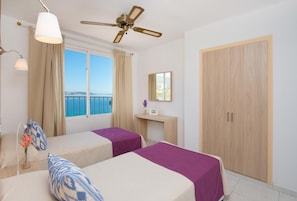 Habitación de hotel con dos camas, un ventilador de techo, una ventana con vista al mar, un armario de madera y una mesita con una lámpara.