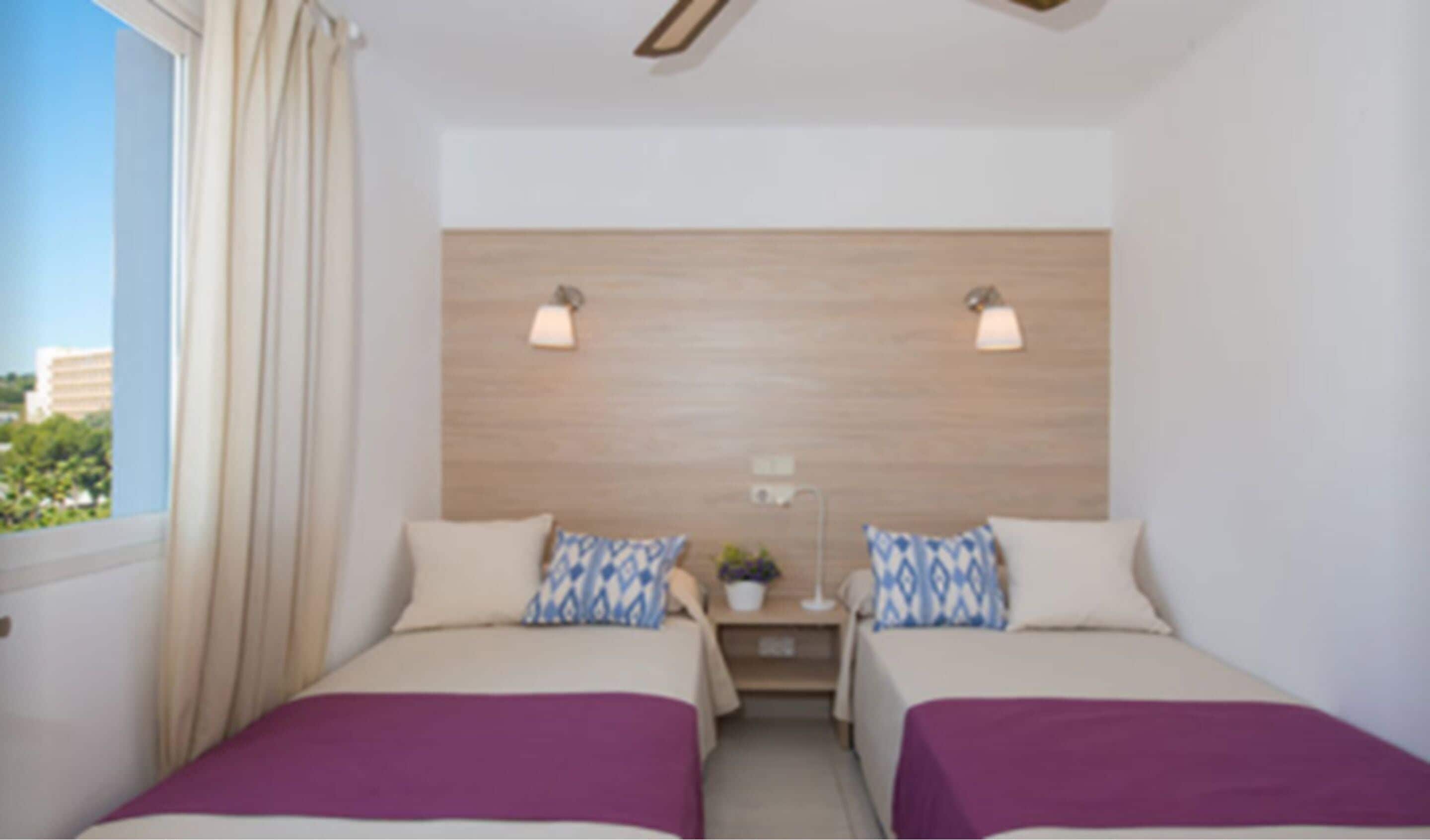 A hotel room with two beds, a wooden headboard, bedside lamps, and a window with curtains.