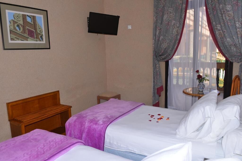 Une chambre d’hôtel avec deux lits, une tête de lit en bois, une petite table avec un vase et une fenêtre avec des rideaux.