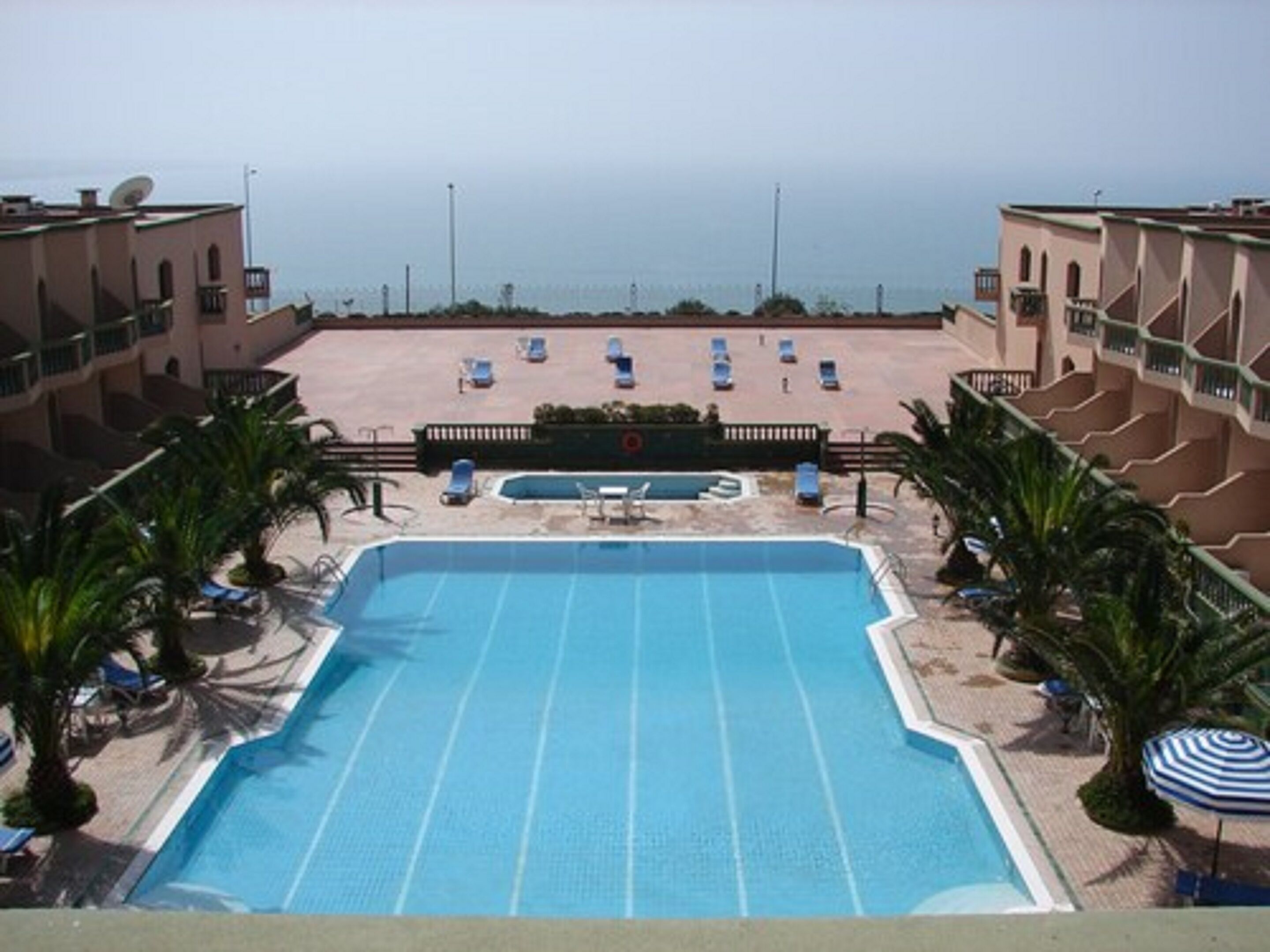 A swimming pool with sun loungers and palm trees, surrounded by a building with balconies.