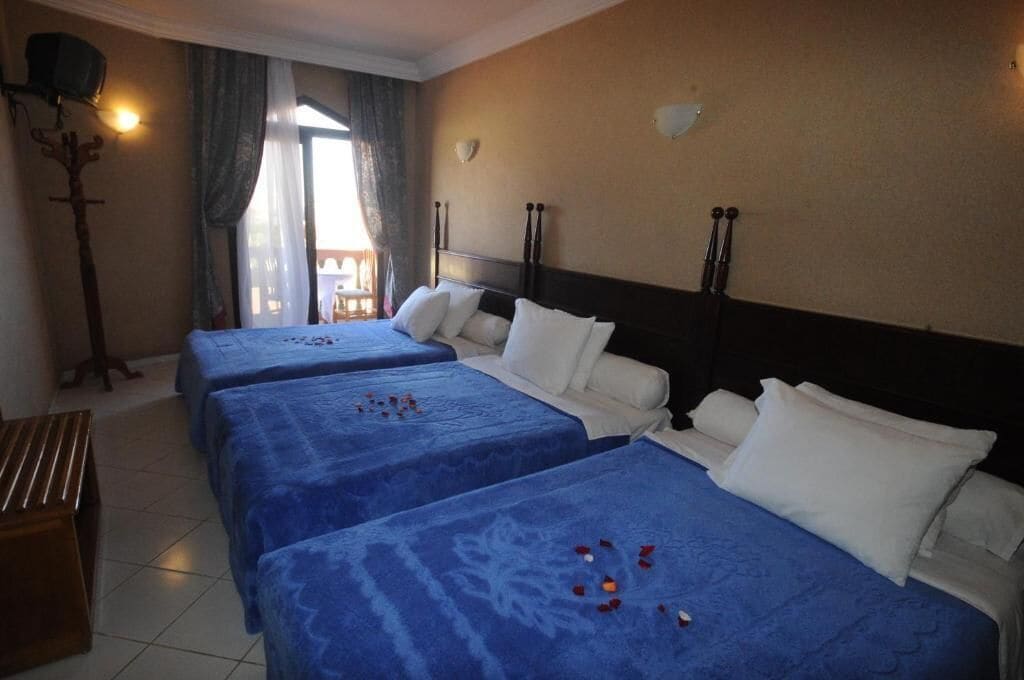 Habitación de hotel con tres camas, cada una con cubrecama azul y almohadas blancas. Hay un banco de madera y una lámpara de pared.