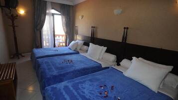 Habitación de hotel con tres camas, cada una con cubrecama azul y almohadas blancas. Hay un banco de madera y una lámpara de pared.