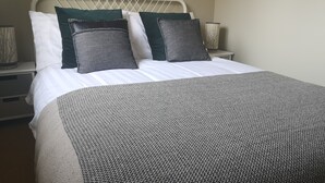 Una cama bien hecha con una colcha a rayas y dos almohadas, una verde y otra estampada, en una habitación sencilla con cabecera blanca y lámparas de noche.