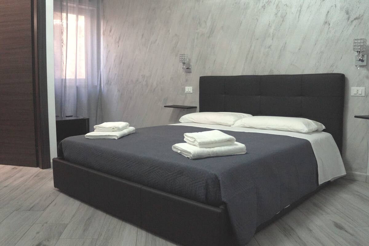 Quarto moderno com cabeceira escura, cama com roupa de cama branca e uma pequena mesa de cabeceira.