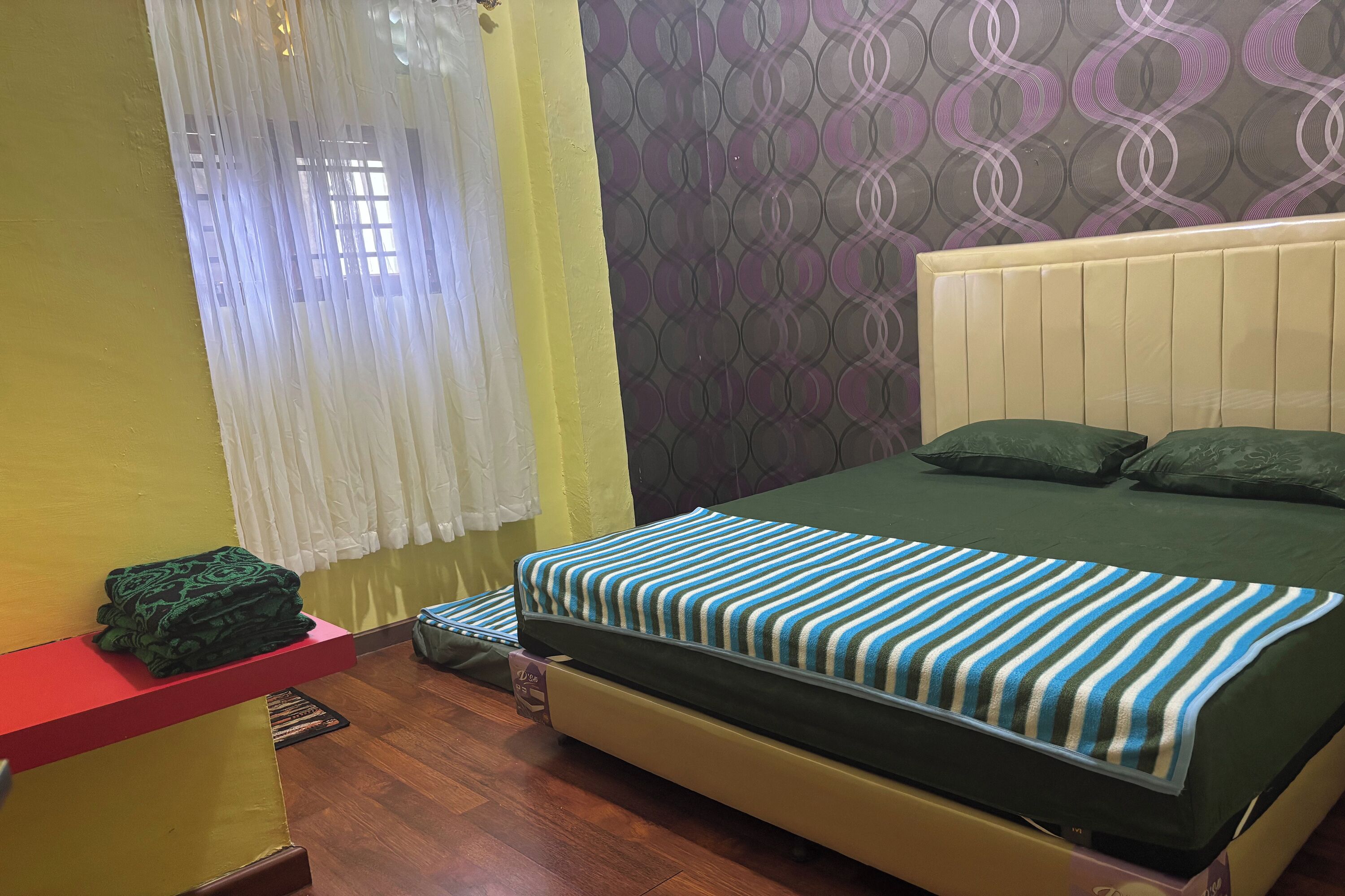 A bed with a striped blanket, a red shelf with a green item, and a patterned wall.