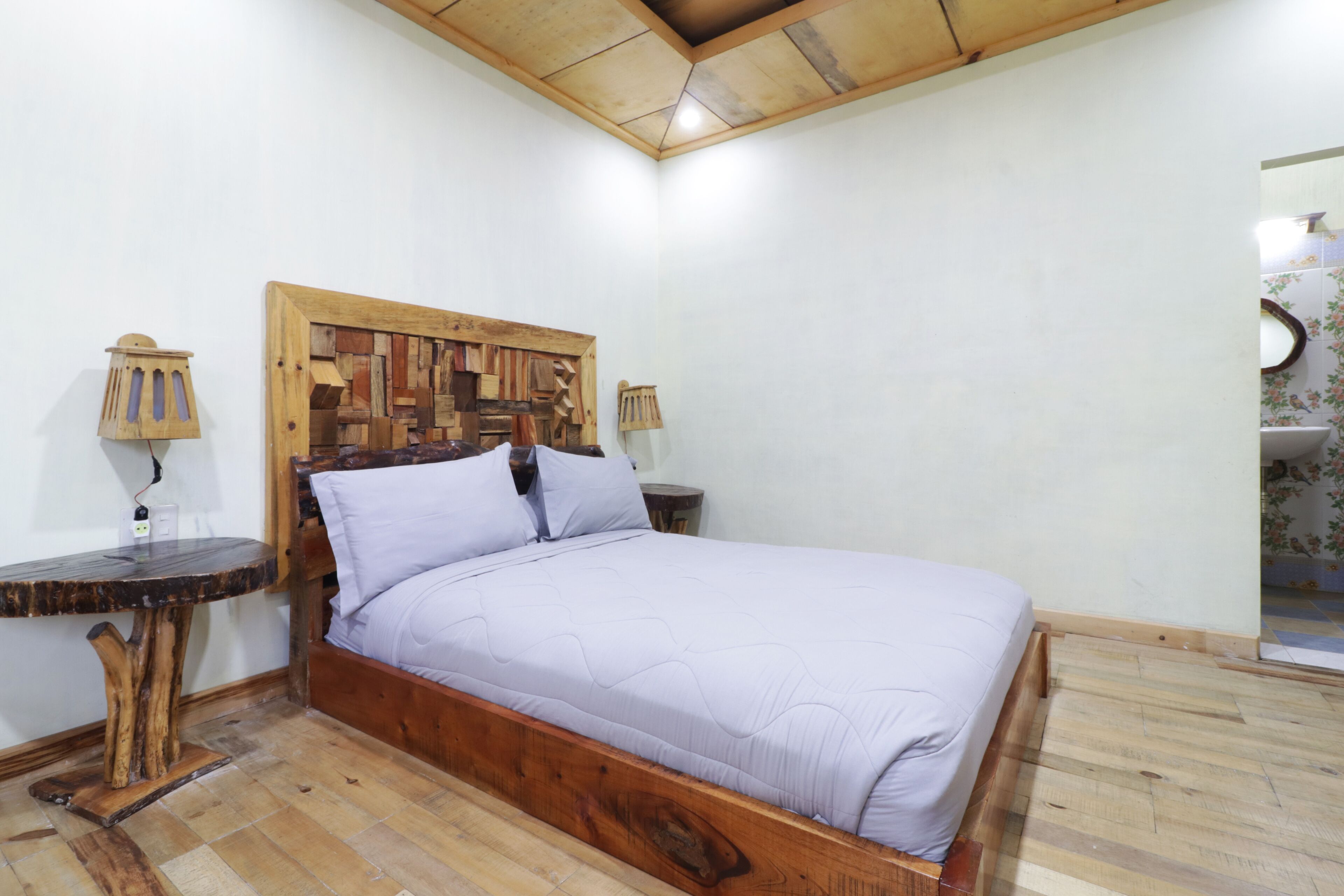 A bedroom with a wooden bed frame, a headboard with a geometric pattern, a round table with a lamp, and a small bedside table.