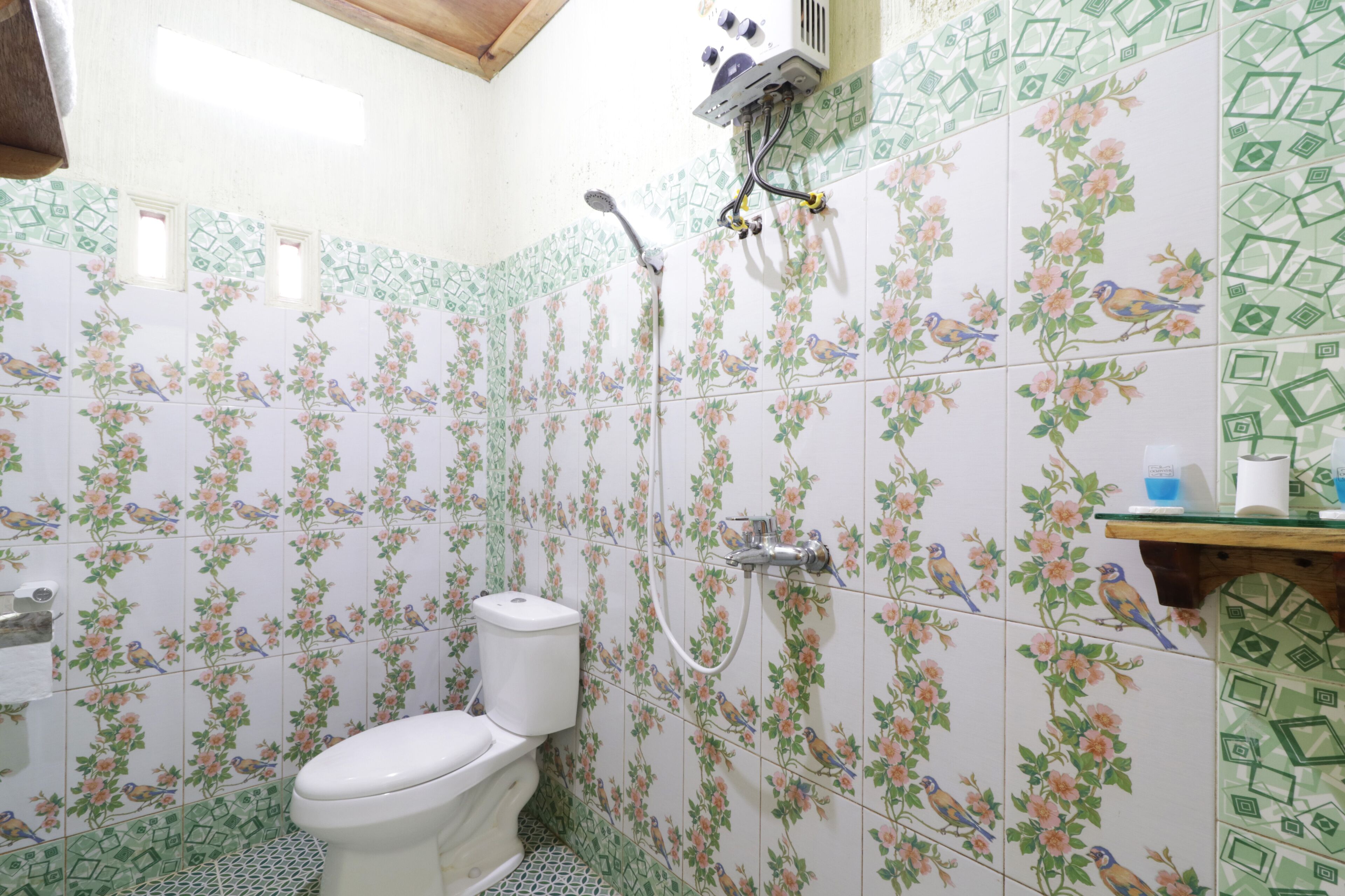 Un baño con patrones de azulejos florales, un inodoro, una ducha y un estante pequeño con artículos de aseo.