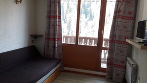 Ein kleines Zimmer mit einem Einzelbett, einem Fernseher auf einem Schrank und einem Fenster mit gemusterten Vorhängen.