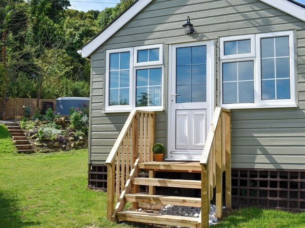 A wooden house with a front porch, a door, and a window with a view of the garden.