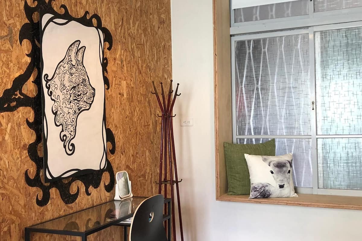 A room with a wooden wall, a framed artwork of a llama, a desk with a chair, a standing lamp, and a window with a view of a building.