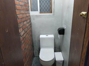 A small, narrow bathroom with a toilet, a trash bin, and a window with a metal screen. - Shadow House (Tainan)
