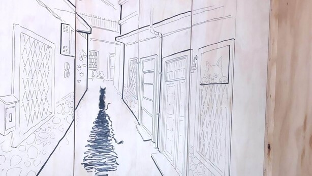 A room with a large wall mural depicting a street scene, a wooden table, two stools, and a fan. - Shadow House (Tainan)
