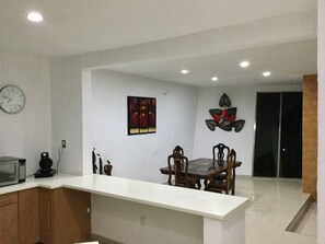 A modern kitchen with a white countertop, wooden cabinets, a dining table with chairs, a clock, and wall art.