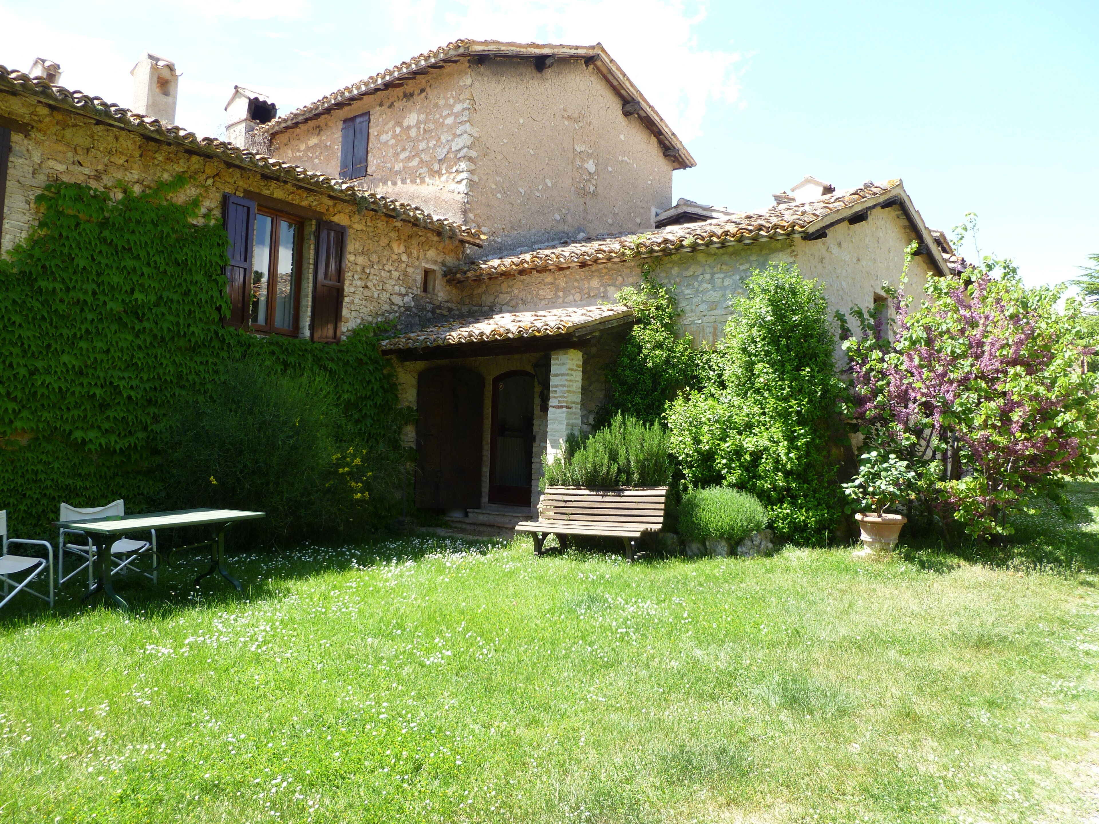 A rustic stone house with a garden, a wooden bench, and a table with chairs.