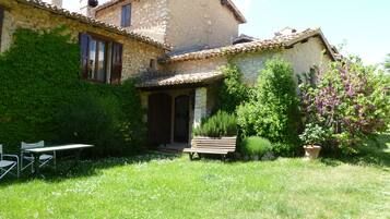 A rustic stone house with a garden, a wooden bench, and a table with chairs.