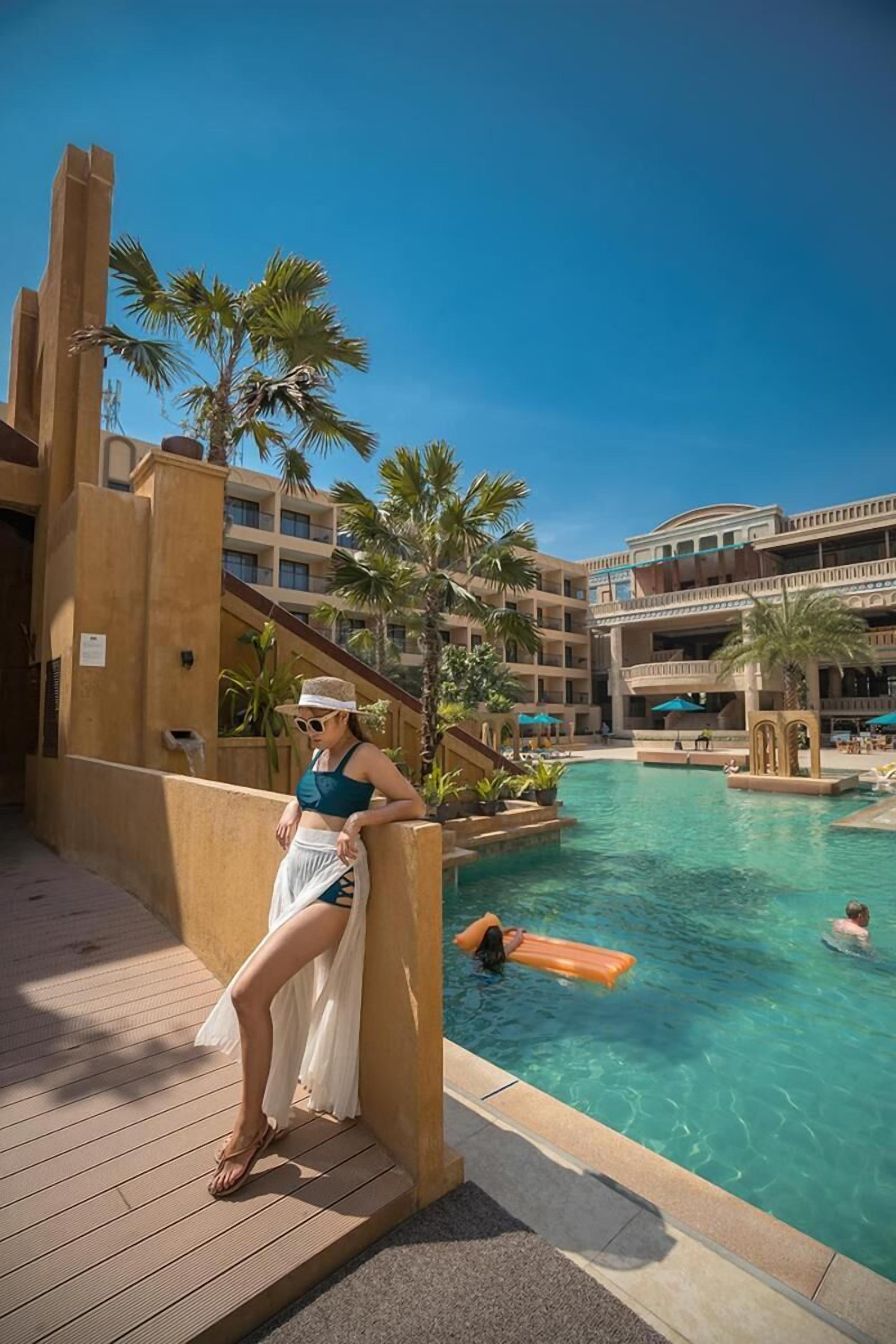 A resort pool area with a person leaning on a railing, palm trees, and a multi-story building in the background.