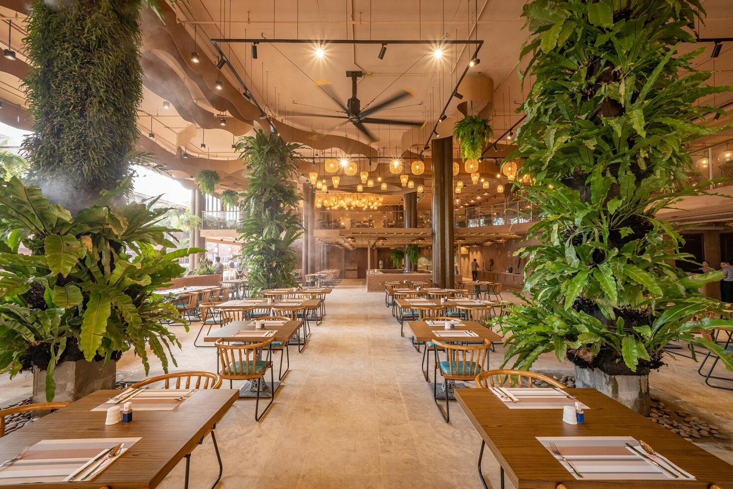 A spacious indoor dining area with multiple tables and chairs, large potted plants, and hanging pendant lights.