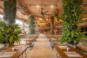 A spacious indoor dining area with multiple tables and chairs, large potted plants, and hanging pendant lights. - Pamookkoo Resort (Karon)