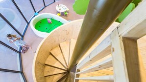 A spiral staircase with a wooden finish, surrounded by glass walls and a green play area with a table and chairs.