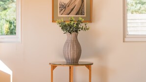 Un jarrón con follaje verde sobre una mesa redonda de madera, frente a una pared con una obra de arte enmarcada.