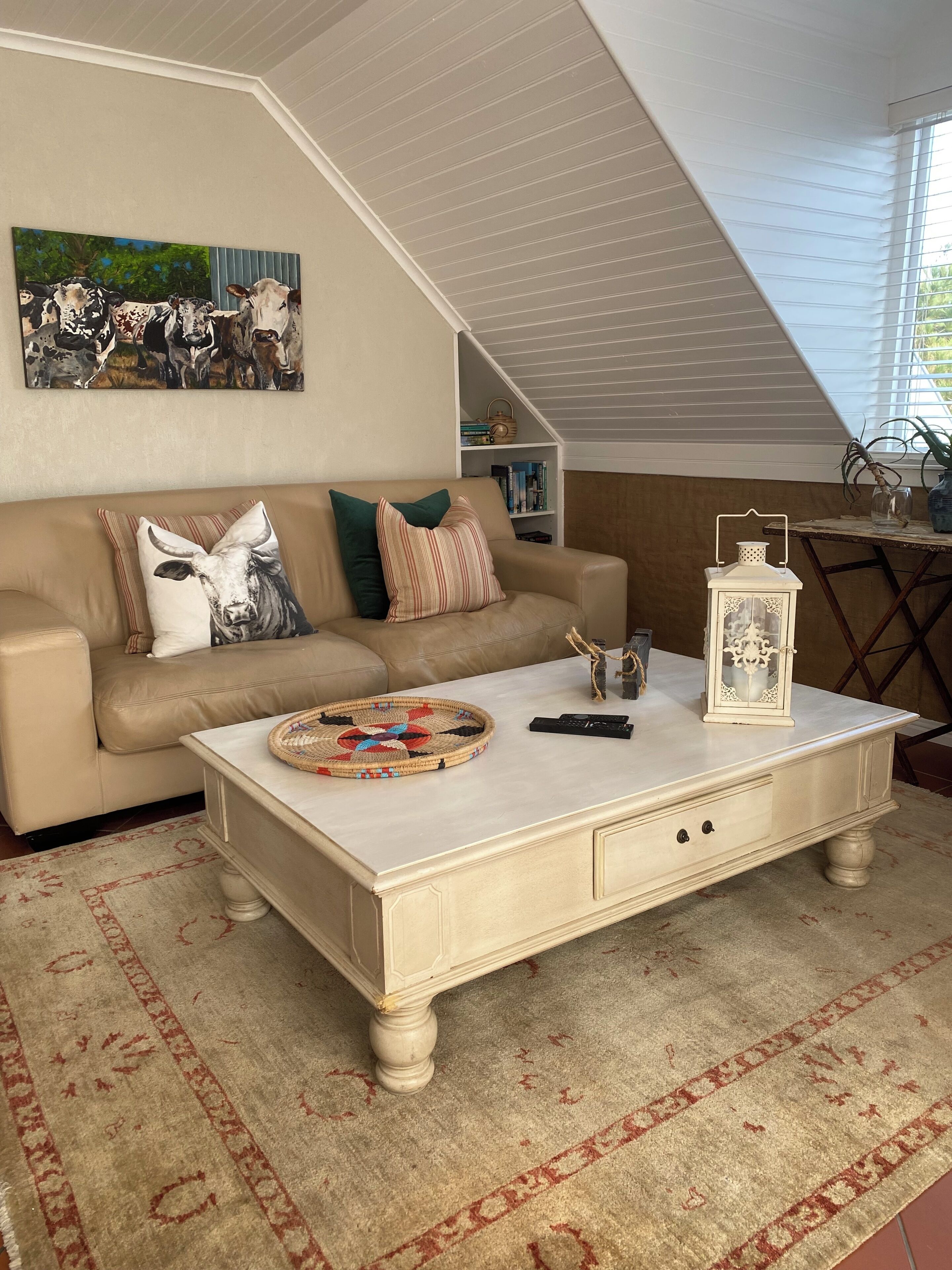 Una sala de estar con un sofá beige, una mesa de centro blanca, un tapete estampado y un cuadro con vacas en la pared.