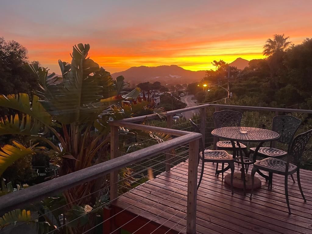 Terraza de madera con una mesa y sillas, con vista a un atardecer y montañas al fondo.