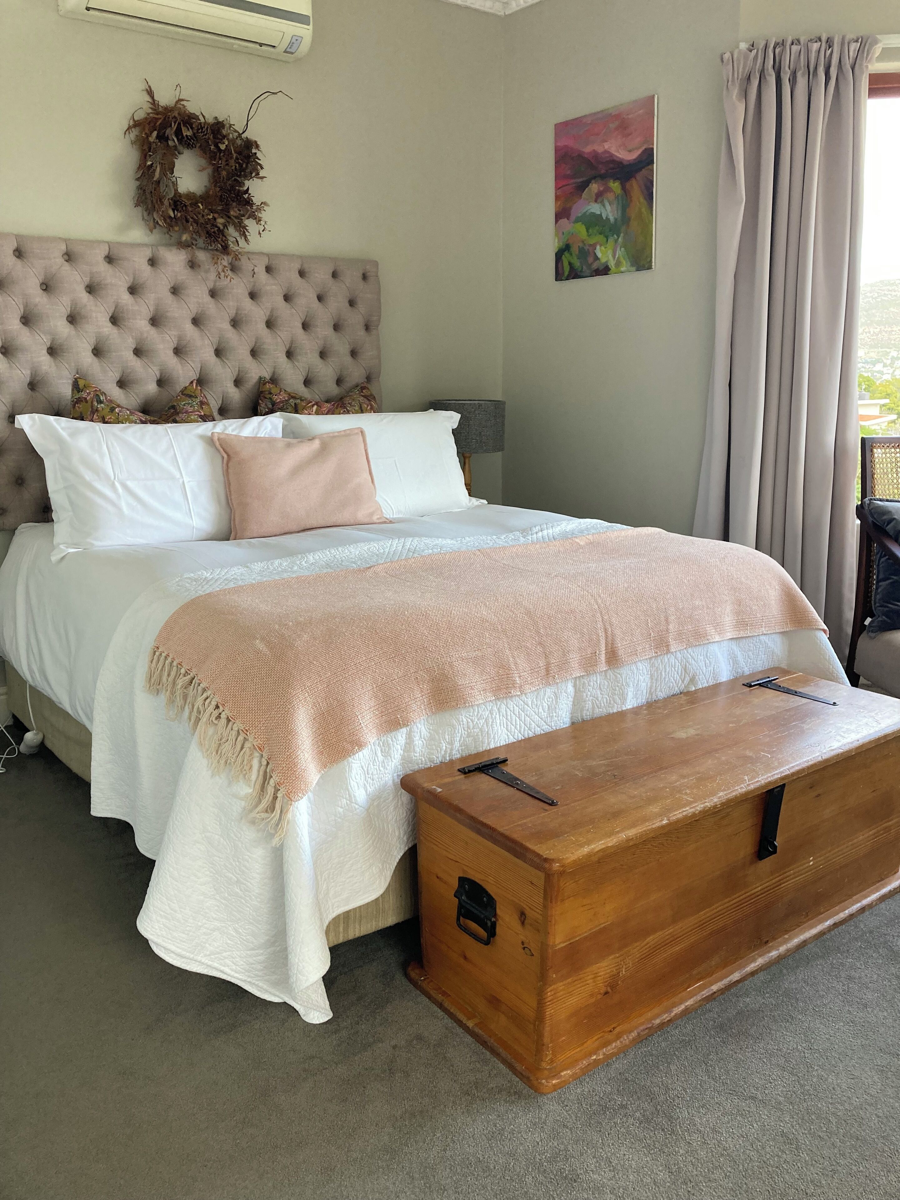 A bedroom with a tufted headboard, a bed with white and peach bedding, a wooden chest, a wall-mounted air conditioner, a wreath, and a painting.