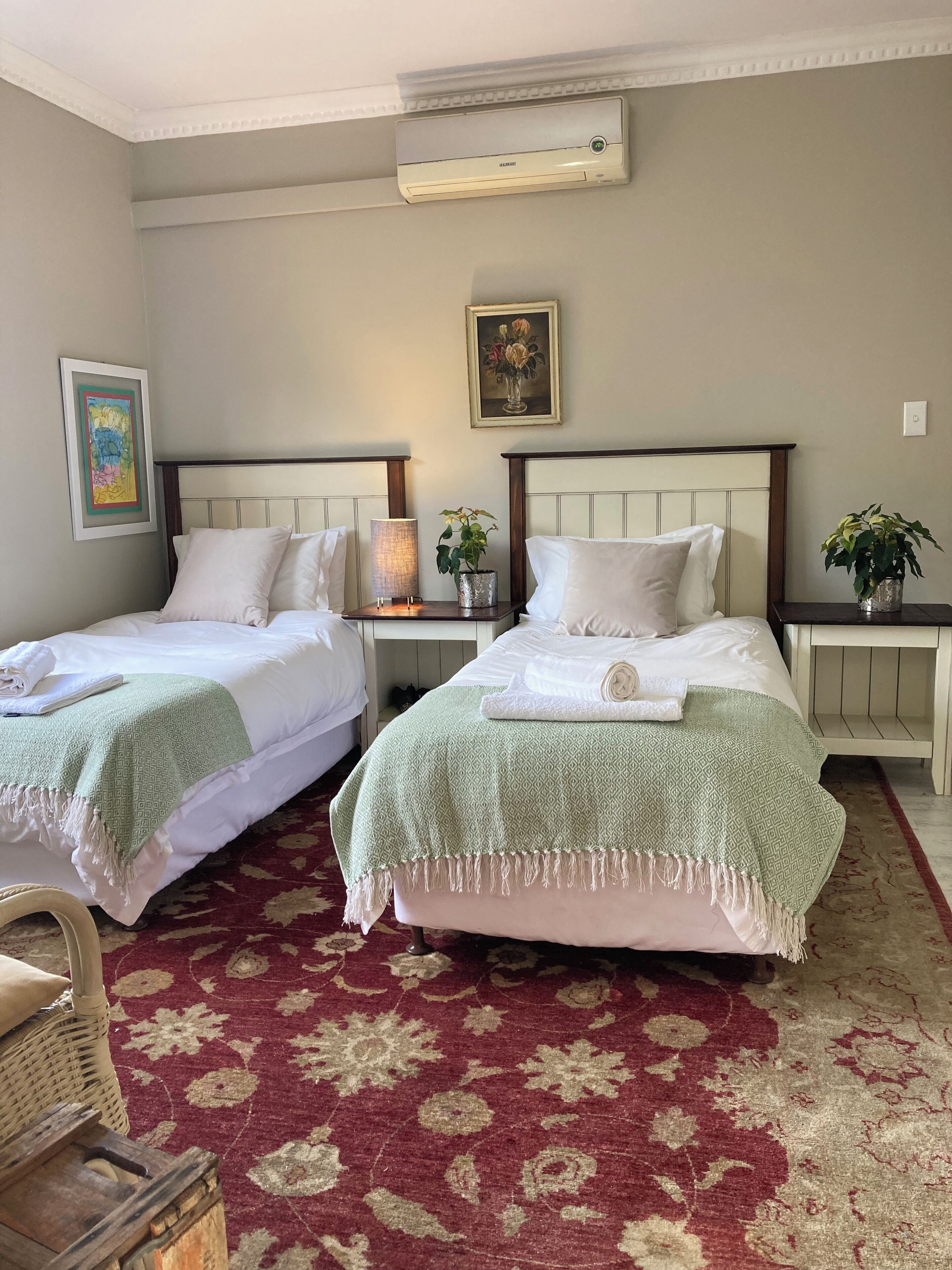 A hotel room with two beds, a red patterned carpet, a wall-mounted air conditioner, a framed painting, and a bedside lamp.
