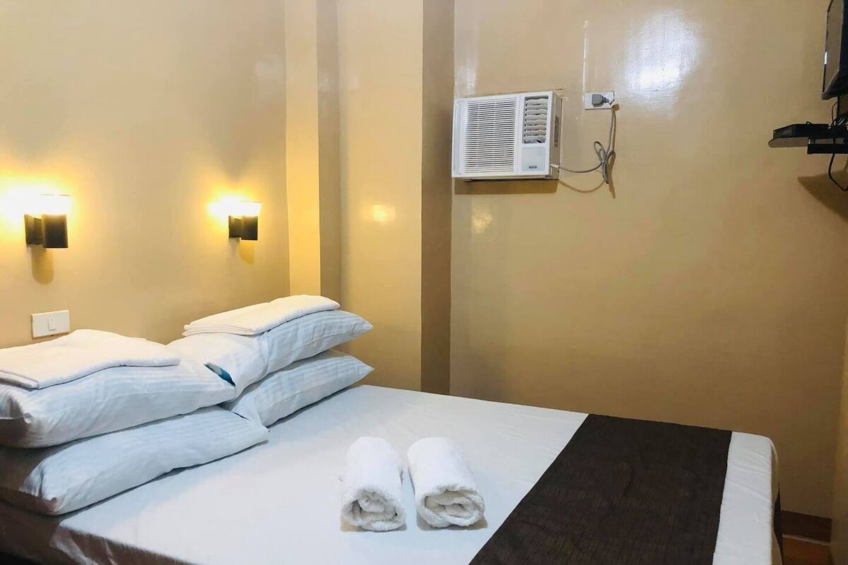 A hotel room with a bed, two wall-mounted lights, an air conditioning unit, and a power outlet.