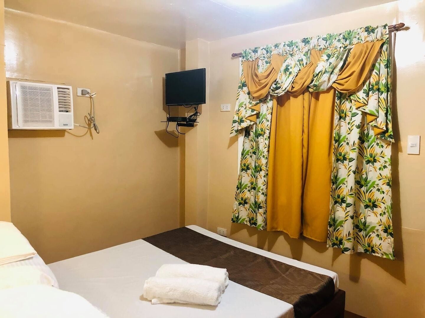 A hotel room with a bed, a TV mounted on the wall, an air conditioning unit, and curtains with a floral pattern.