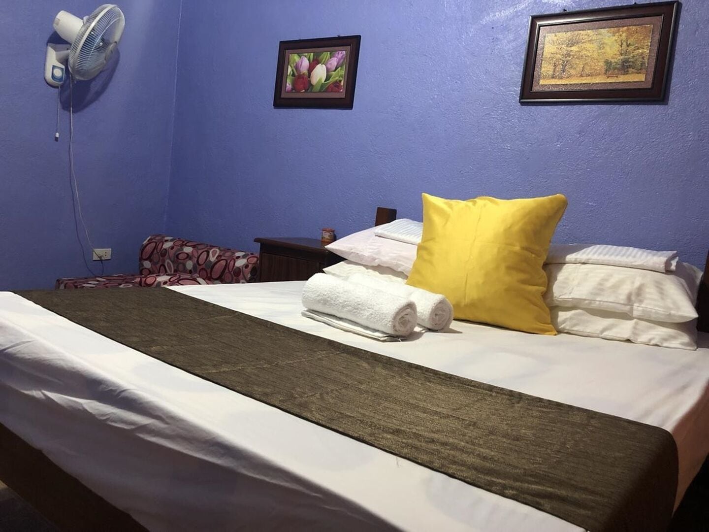 Una habitación de hotel con una cama, un ventilador, dos cuadros enmarcados y una almohada amarilla.