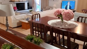 Een eetgedeelte met een houten tafel, stoelen en een bloevas. Achtergrond is een bank en een televisie.