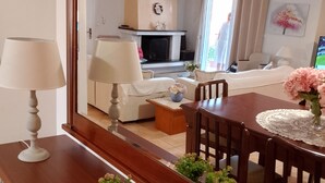 Een eethoek met een houten tafel, stoelen en een spiegel die een woonkamer reflecteert, met een bank, televisie en een open haard.