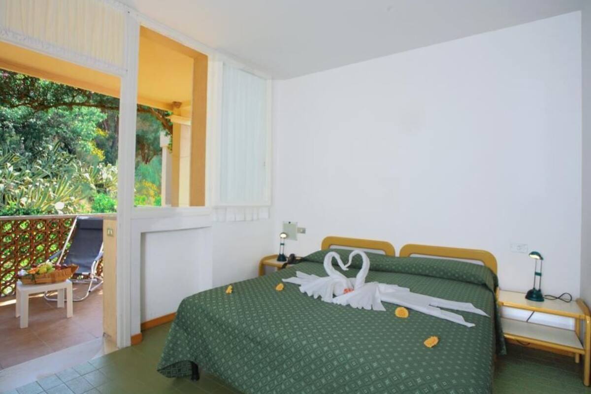 Une chambre avec une couvre-lit à motifs verts, une tête de lit en bois, une table de chevet avec une lampe, et une vue sur un balcon avec une chaise et un panier de fruits.
