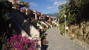 A stone pathway leading to a building with balconies adorned with purple flowers, surrounded by greenery and a mountainous backdrop.
