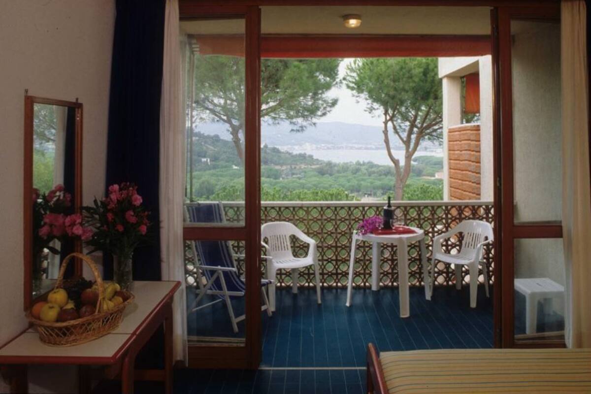 Un balcon avec vue sur un paysage verdoyant et un plan d’eau. On y trouve deux chaises en plastique blanches et une petite table ronde sur laquelle est posé un vase de fleurs. Un panier en osier rempli de fruits se trouve sur une table à gauche.