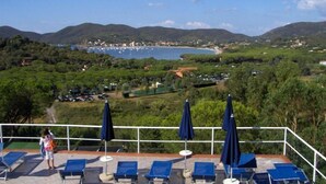 A terrace with blue lounge chairs and umbrellas overlooking a coastal area with hills and a marina.