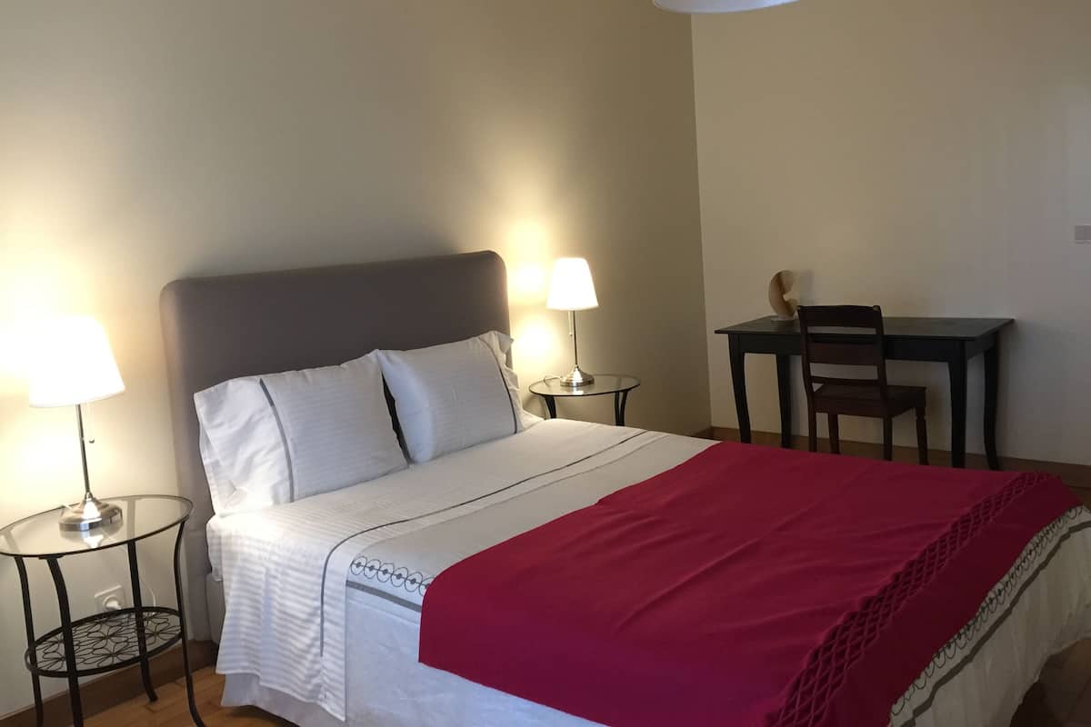 A neatly made bed with white bedding and a red throw blanket, a small round table with a lamp, a chair, and a desk with a chair in a room with plain walls and a ceiling light.