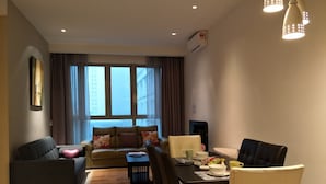 Una sala de estar moderna con sofá, mesa de comedor y sillas. Se ve un ventanal con cortinas, un equipo de aire acondicionado en la pared y un televisor de pantalla plana.