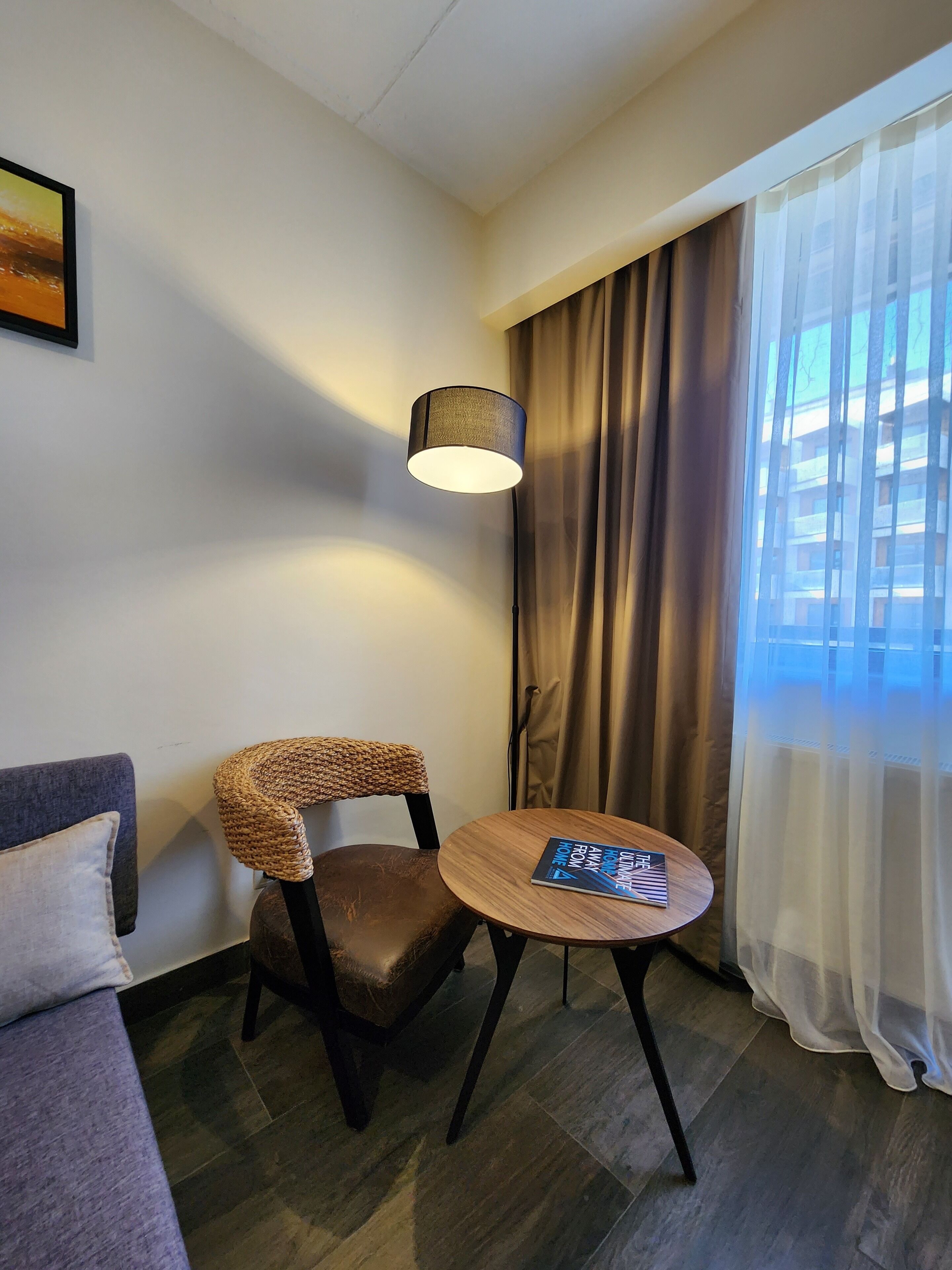 A hotel room with a sofa, a chair with a woven backrest, a small wooden table with a book, a floor lamp, and a window with curtains.