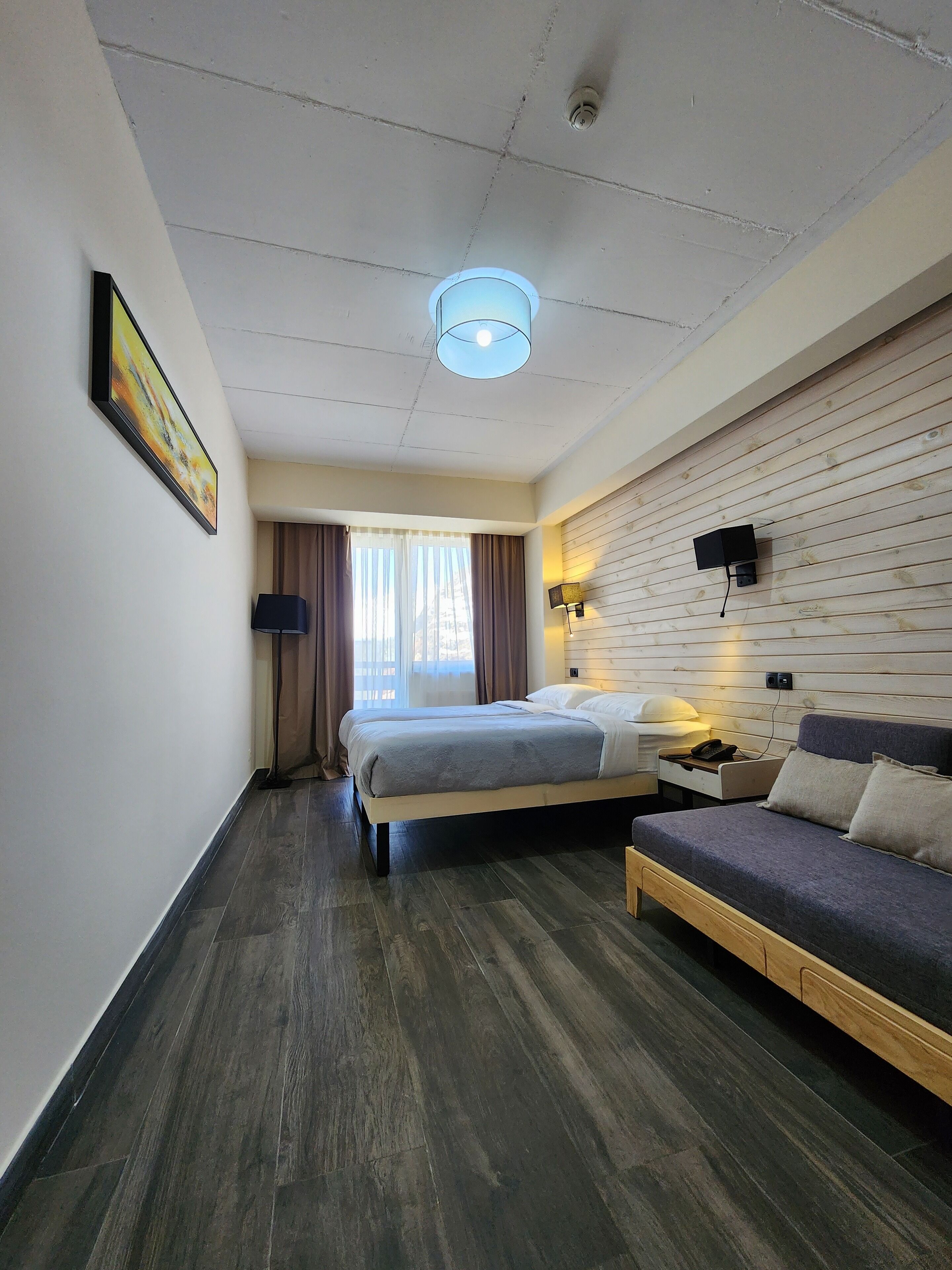 A hotel room with a large bed, a sofa, a desk with a lamp, and a wall-mounted TV.