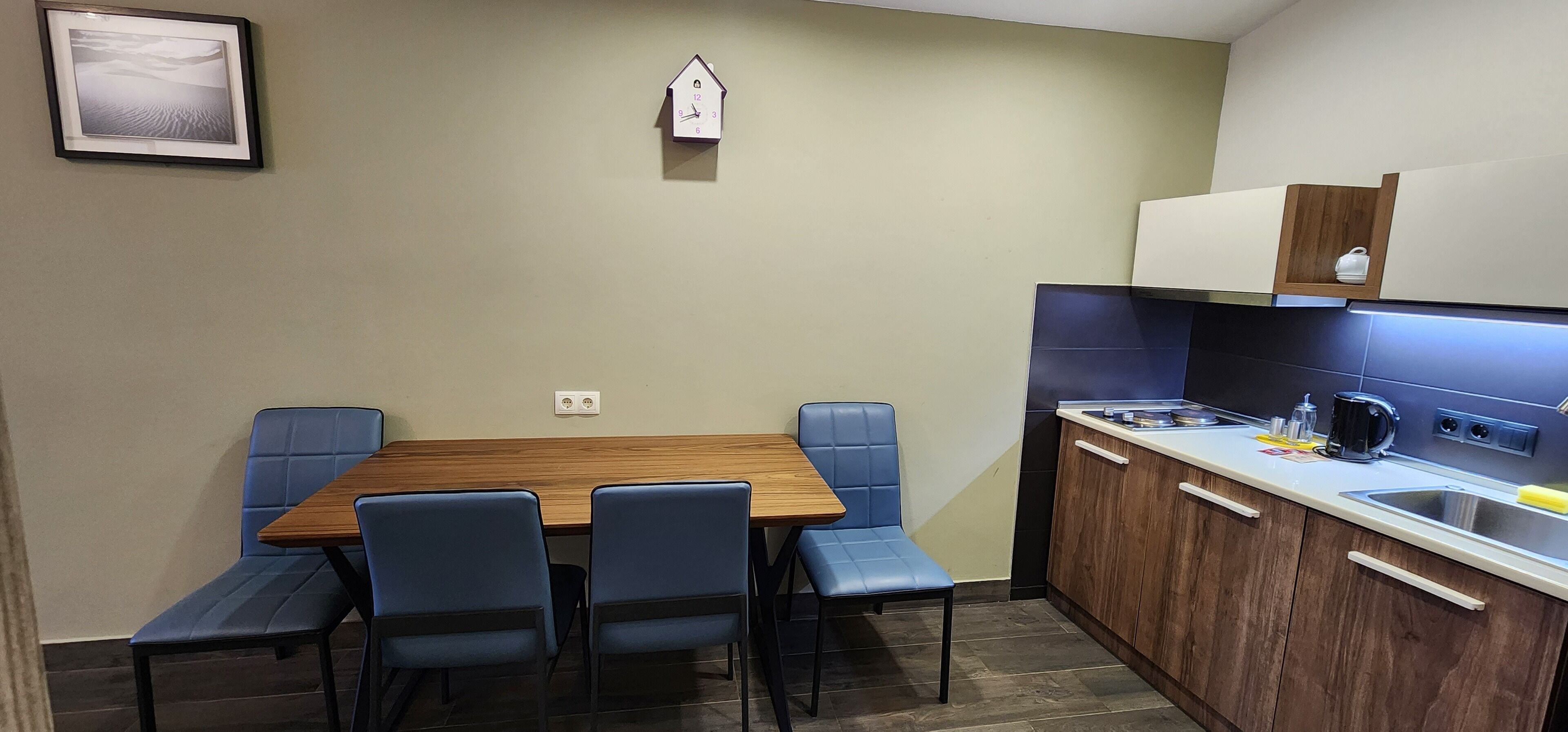 Una cucina compatta con un tavolo di legno e sedie blu, una piccola sveglia al muro e una macchina da caffè sul ripiano.