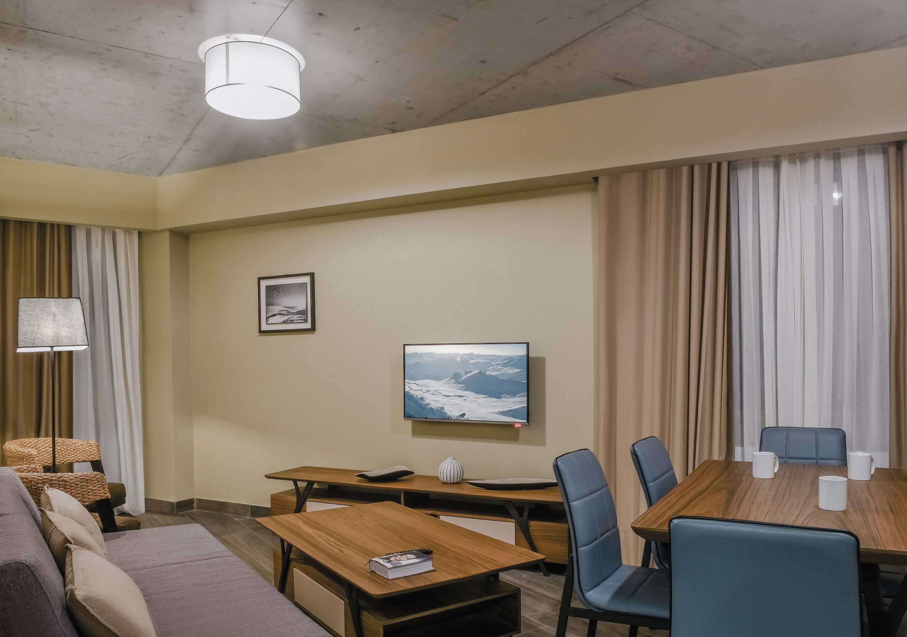 A hotel room with a sofa, a wooden table, a desk, and a dining table with chairs. There is a television mounted on the wall and a picture frame on the wall.