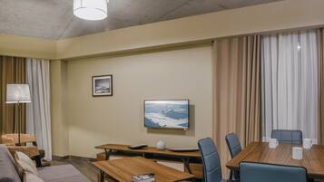 A hotel room with a sofa, a wooden table, a desk, and a dining table with chairs. There is a television mounted on the wall and a picture frame on the wall.
