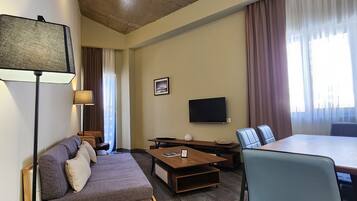 A modern hotel room with a sofa, a dining table, a TV, and a large window with curtains.