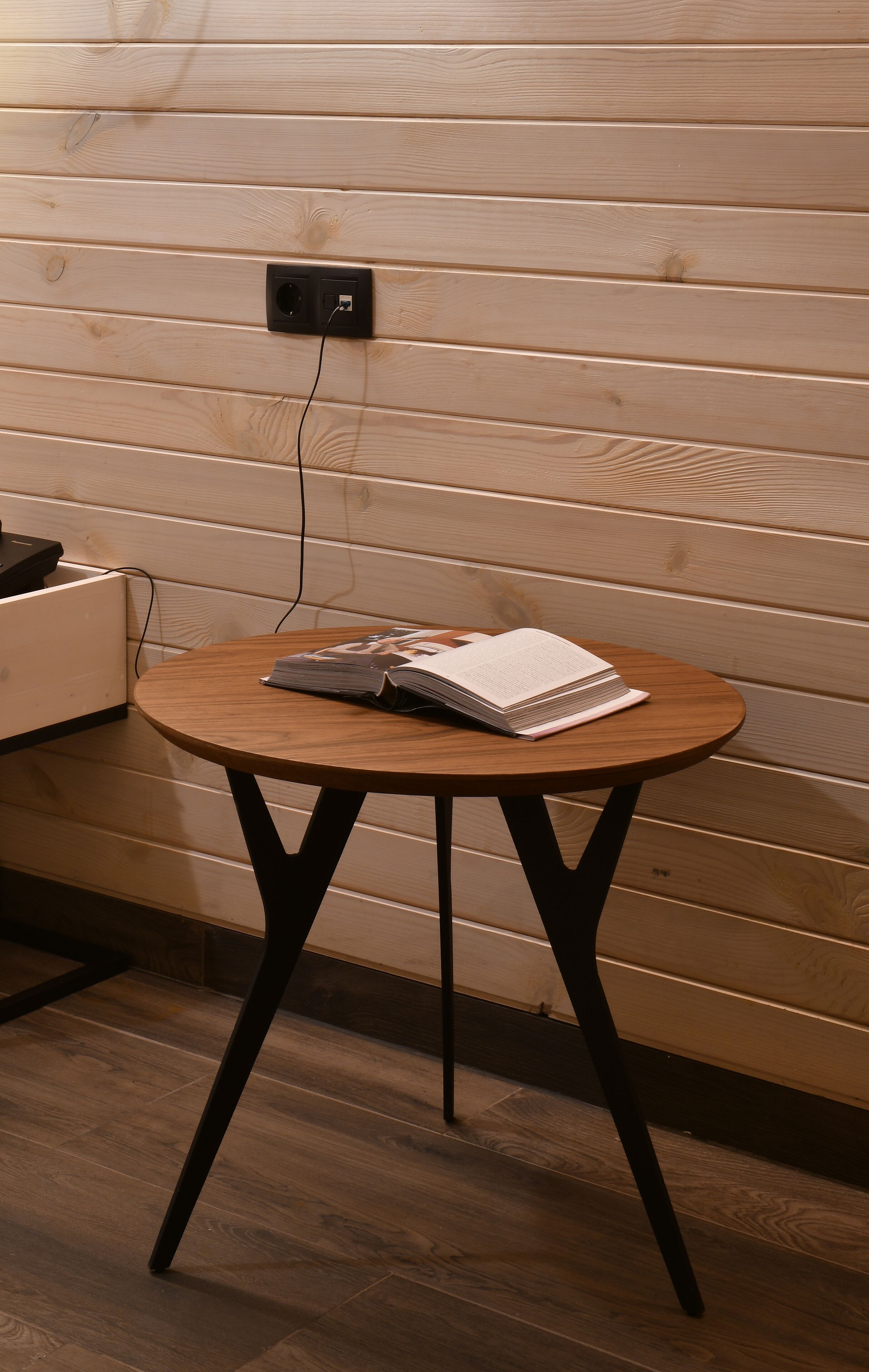 Een houten tafeltje met een notitieboekje erop, geplaatst tegen een houten wand met een stopcontact erboven.